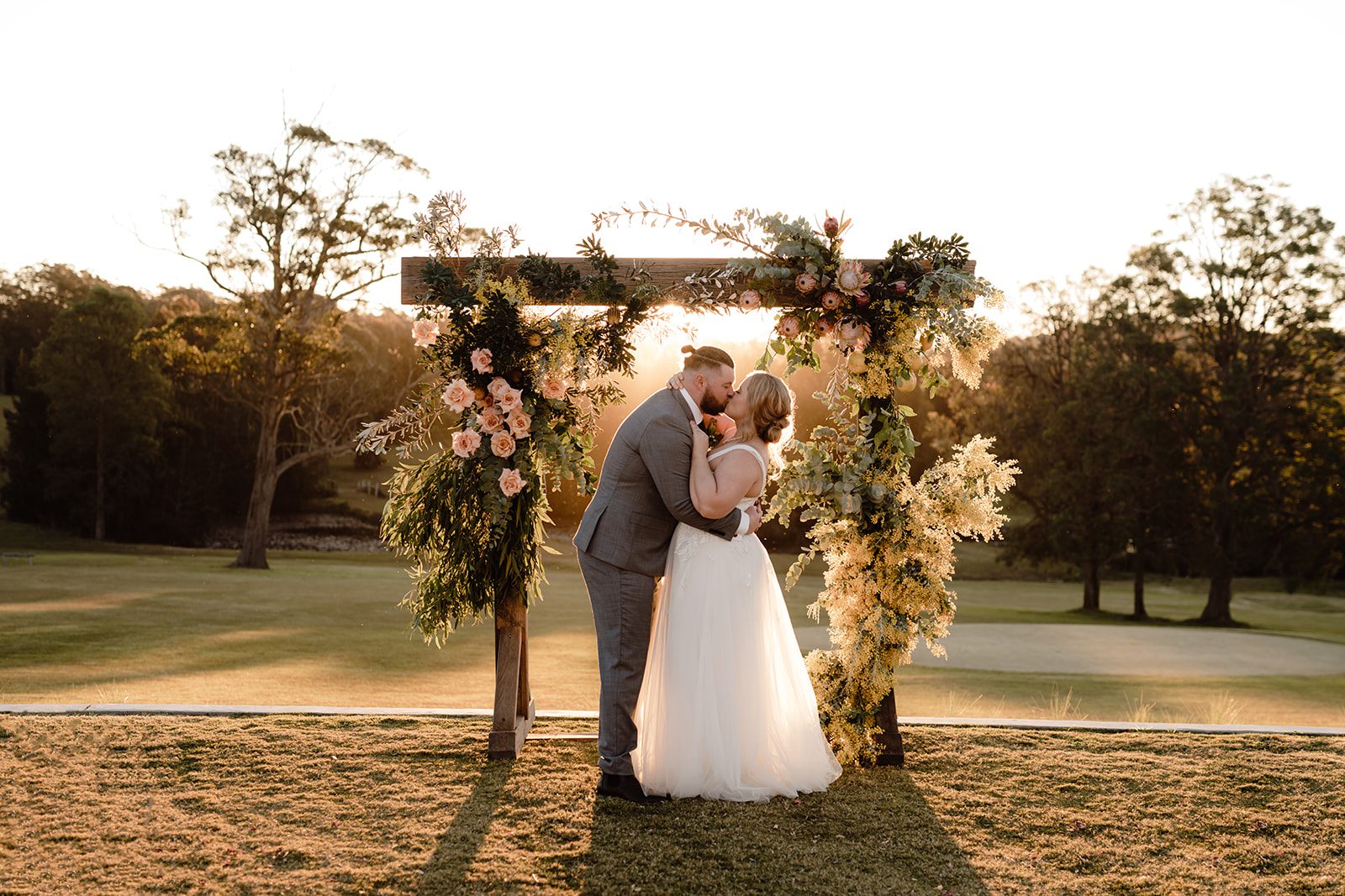 Bride and groom standing infront of floral arbour at the Oaks ranch kissing with a breathtaking sunest