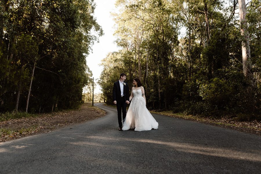 Kayleigh & Michael walking down the road holding hands looking at each other with the side of the roads covered in trees