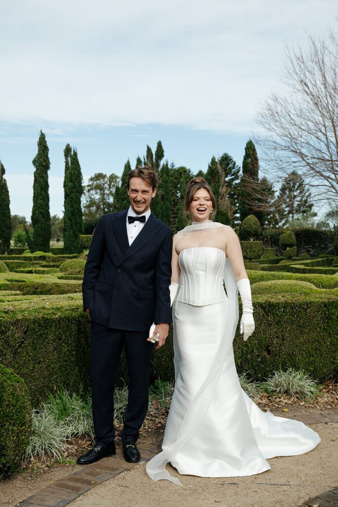 Bride and groom laugh together in front of the topiary maze at The Merribee, with cypress trees and the formal gardens behind them.