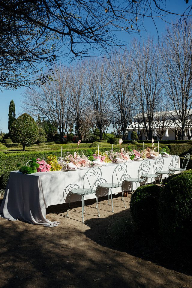 A long wedding reception table set up in the topiary garden at The Merribee, with white linen, pastel florals, and wrought-iron chairs.